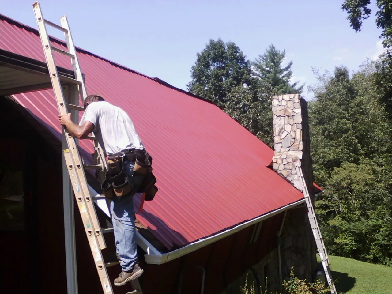 Roofer on a ladder beside a red metal roof and stone chimney — Soffit & Fascia Repair by Roofing Brothers in Dellroy